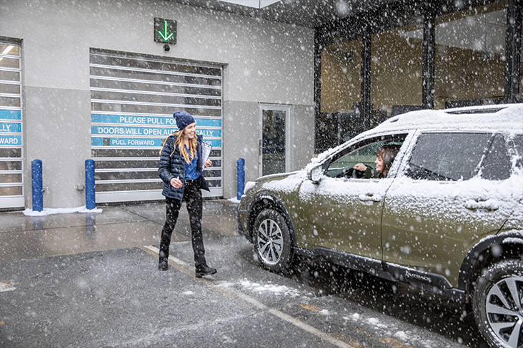 A young woman is walking toward a Subaru vehicle pulled up outside a service area. She is carrying a clip board and smiling. There is another woman in the driver's seat with her window rolled down. It's snowing outside, and the Subaru is covered in snow.