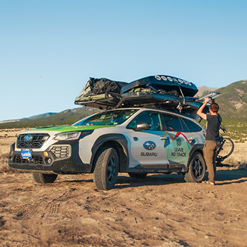 A Subaru Outback Wilderness Leave No Trace vehicle is parked on a clearing with mountains in the background. A male is loading an item on top of the vehicle's roof, and the roof is loaded with a hard-shell carrier and camping equipment.