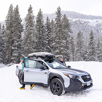 Two ladies are exiting a silver Subaru Outback Wilderness parked on a snowy hill with tall evergreen trees dappled with snow in the background.