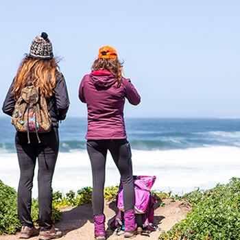 Two women stand on a cliff with their backs to the camera looking out onto the ocean. They are both dressed in hiking boots, pants, jackets, hats and backpacks.