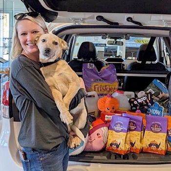 Carrie Cahoon is standing in front of a Subaru that's loaded with pet supplies at an event. She's holding her Australian cattle dog, Maverick, in her arms and is smiling.