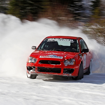 A student learning to drift through the snow in a Subaru Impreza. The red Impreza stands out against the white snow, and snow is being kicked up into the air by its rear wheels.