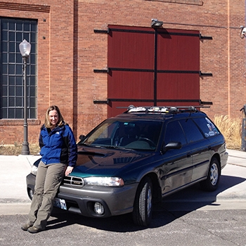 Amanda Isaacs in Wyoming with her 1997 Subaru Outback, Mr. Lizard, several months after her road trip. She is leaning back on the hood of the vehicle, wearing brown slacks and a blue, long-sleeved jacket. A brick building is behind the Outback. 