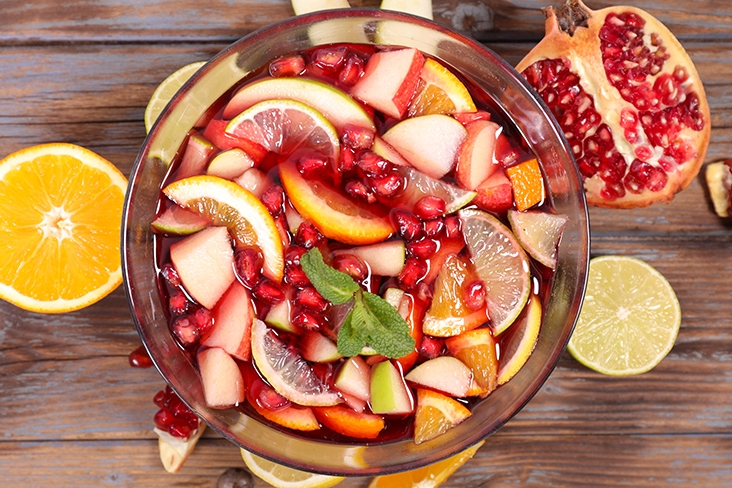 Large glass punch bowl filled with freshly sliced fruit, including lemons, pomegranate seeds, and oranges, and red punch.