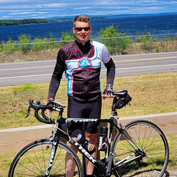 Jack Frost, who is participating in the Trek Across Maine fundraiser, stands behind a bike wearing active gear. A road and large body of water are behind him.