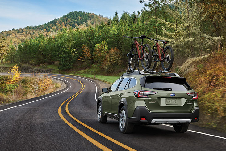 A 2025 Subaru Outback Touring in Autumn Green Metallic is driving on a two-lane road with two bikes on the roof.