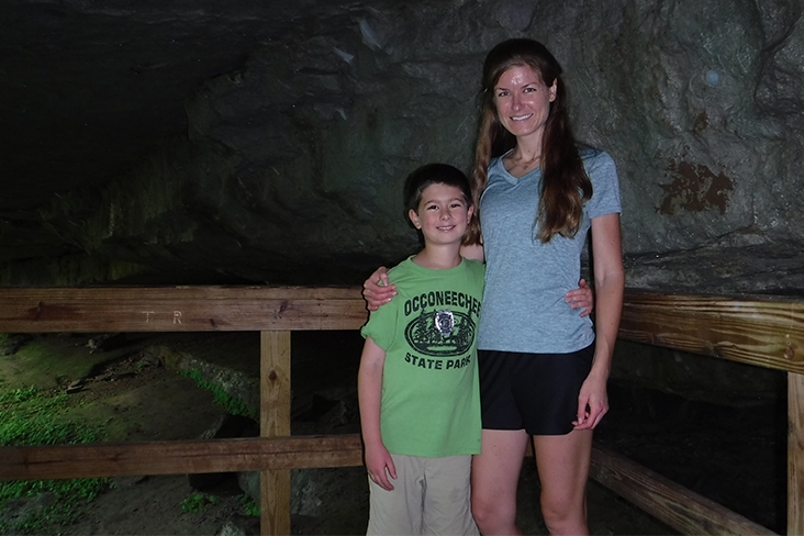 Casey Amarnek and her son, Caden, at Mammoth Cave National Park in Kentucky. They are both smiling and standing against a wooden rail; Casey has her arm around Caden’s shoulder. Dark cave walls can be seen behind them.