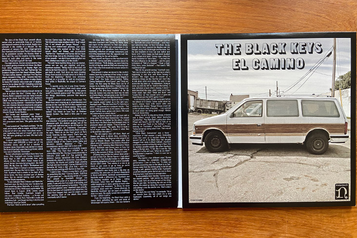 A shot of the El Camino album by The Black Keys. It has a gatefold style cover, which is open and lying flat on a tabletop. On the left side, words take up the entire side. On the right, it says The Black Keys, El Camino, and underneath the words is an image of a Grand Voyager vehicle. 