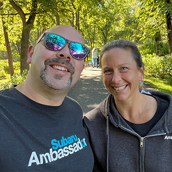 Kyle and Stacey Williams are taking a selfie on a walking path that is surrounded by deciduous trees. They are both smiling. 