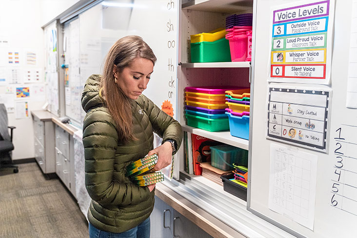 Kimber Cross standing in front of an open cabinet with colorful bins as she organizes supplies in a classroom.
