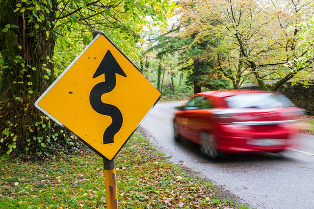 Red,Car,Driving,Past,Dangerous,Winding,Road,Sign,In,A Red,Car,Driving,Past,Dangerous,Winding,Road,Sign,In,A