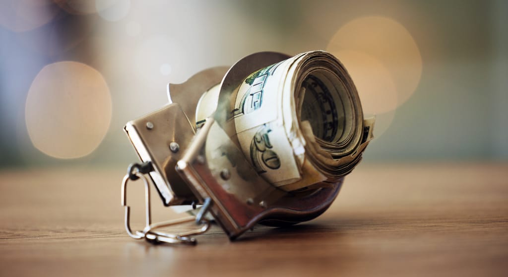 A photograph of a roll of dollar bills inside a pair of handcuffs, on a wooden tabletop A photograph of a roll of dollar bills inside a pair of handcuffs, on a wooden tabletop