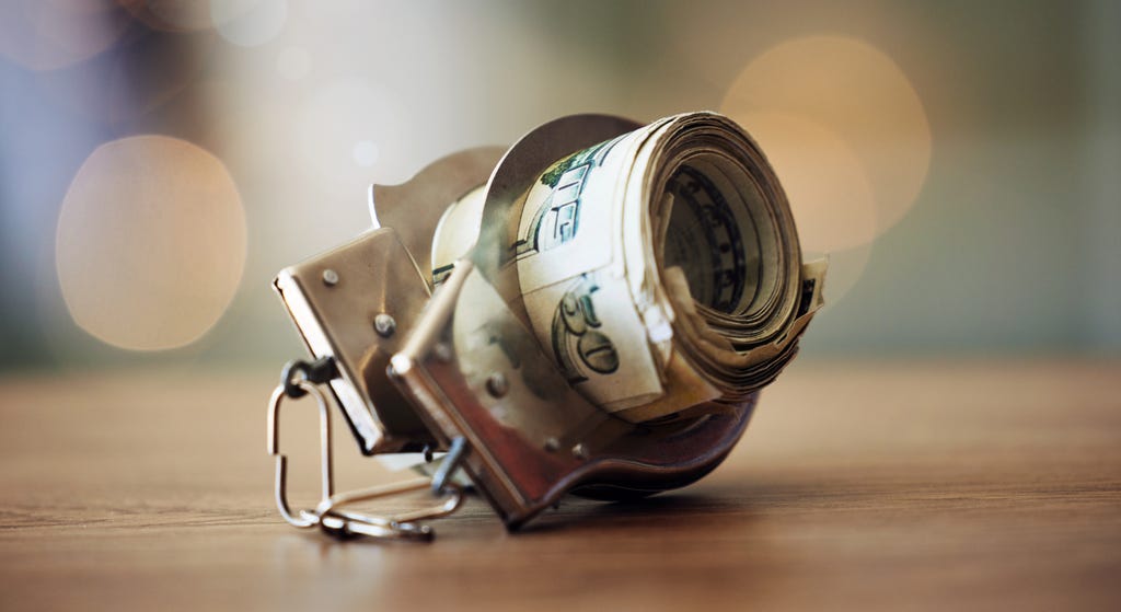 A photograph of a roll of dollar bills inside a pair of handcuffs, on a wooden tabletop A photograph of a roll of dollar bills inside a pair of handcuffs, on a wooden tabletop