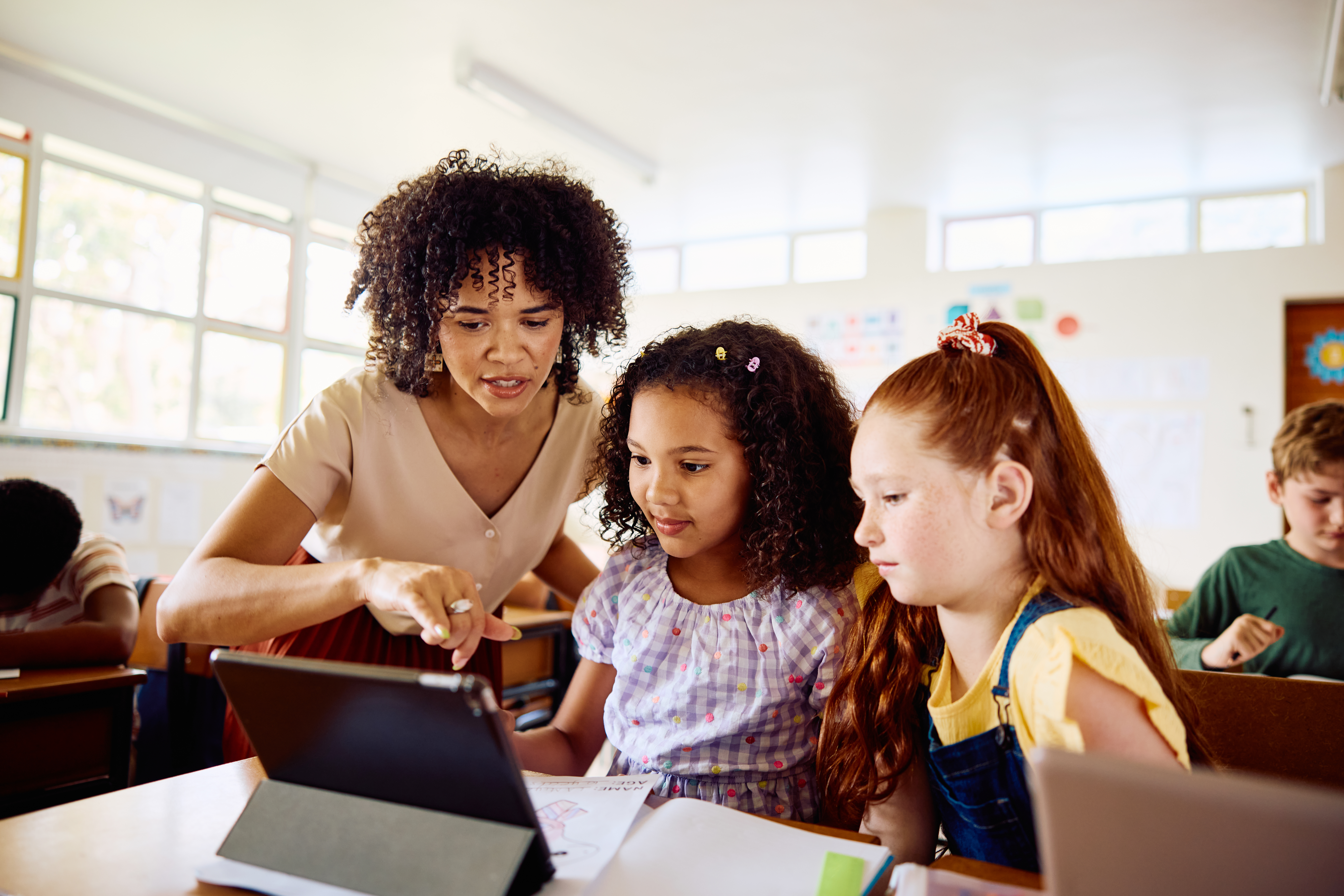 A female teacher in a classroom looks over the shoulders of two young students as they use an iPad as a learning resource.