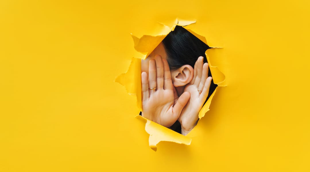 A person, face obscured, behind a sheet of yellow paper. A torn gap in the paper shows the person putting both hands to their ear as though trying to hear something.