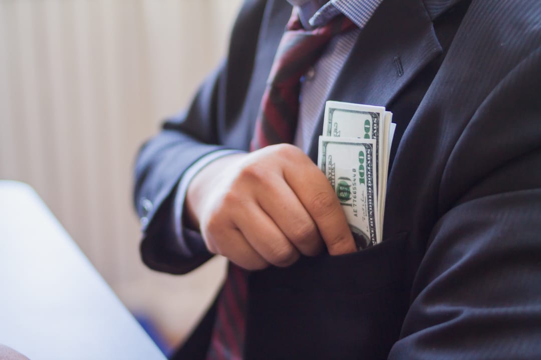 A photograph showing a person in a suit and tie tucking a sheaf of dollar bills into their top pocket