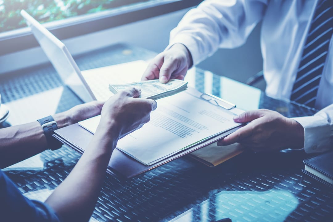 A photo showing a person handing over a sheaf of dollar bills to someone over a desk. The recipient is wearing a shirt and tie. In return, the recipient is handing the other person a clipboard with sheets of paper on it