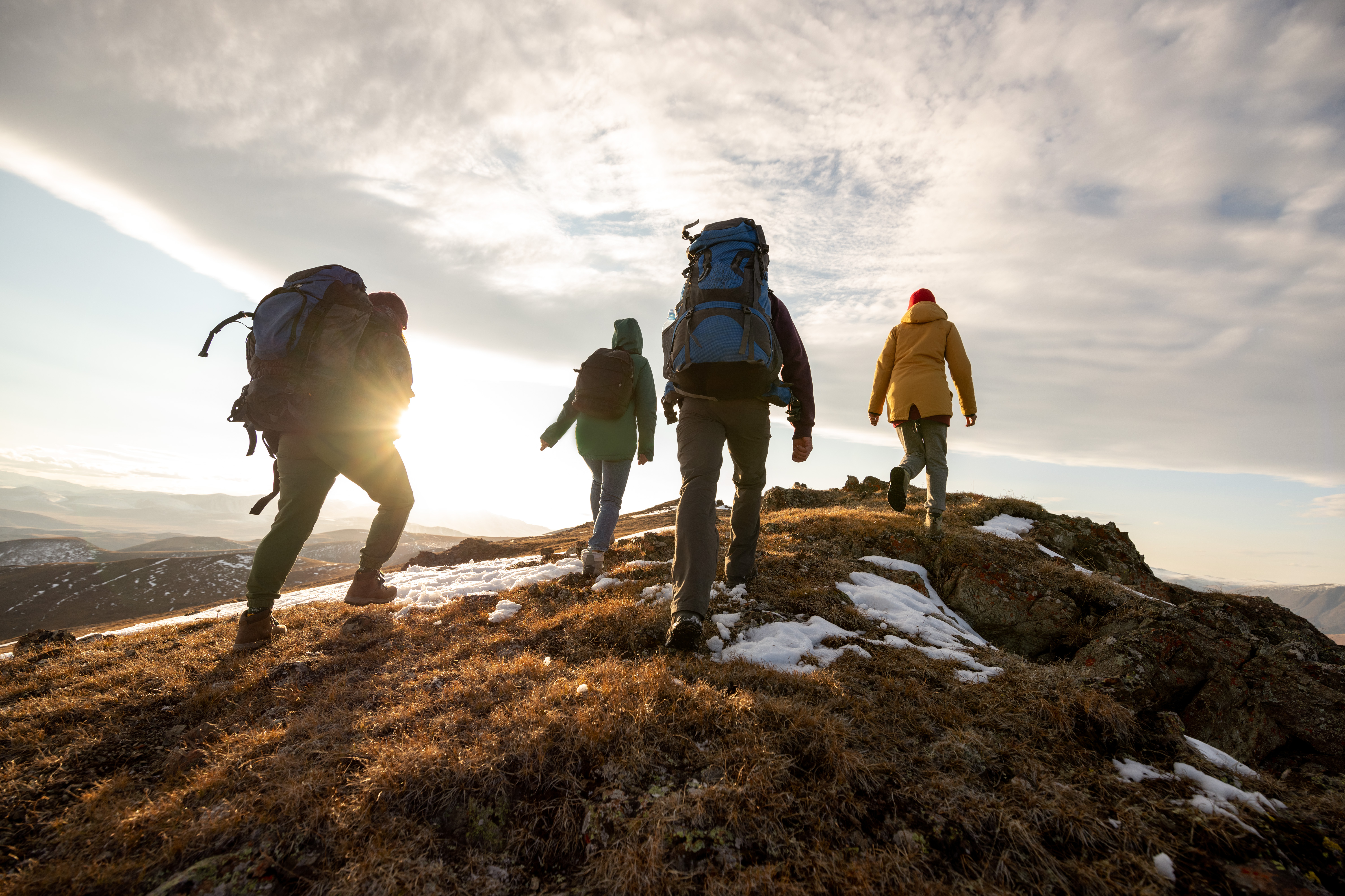 A group of four people with hiking equipment walking up a hill or mountain, taken from behind, during sunset on a cloudy day