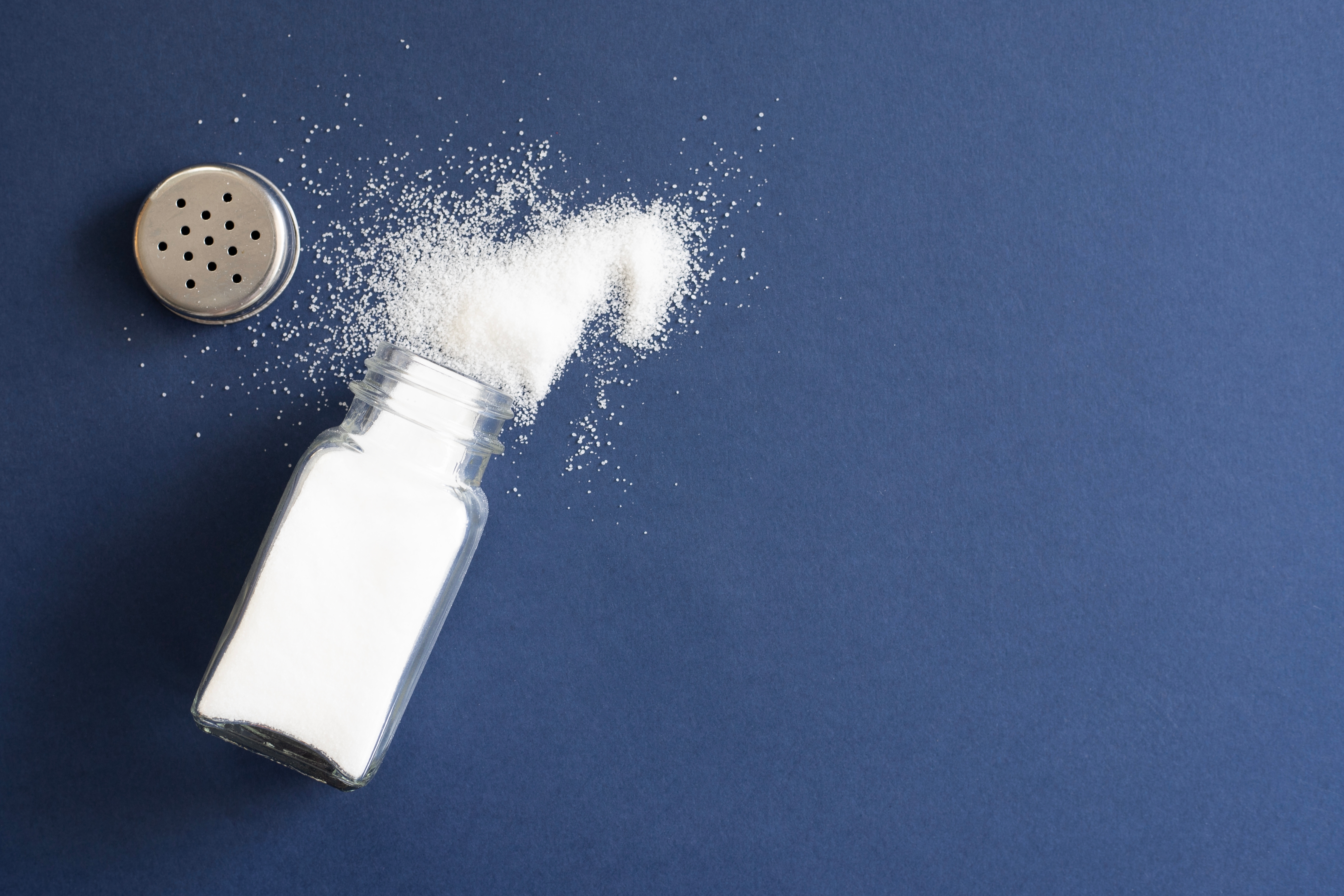 A salt shaker lying flat on a blue tabletop, with the lid off and salt spilling out on to the table