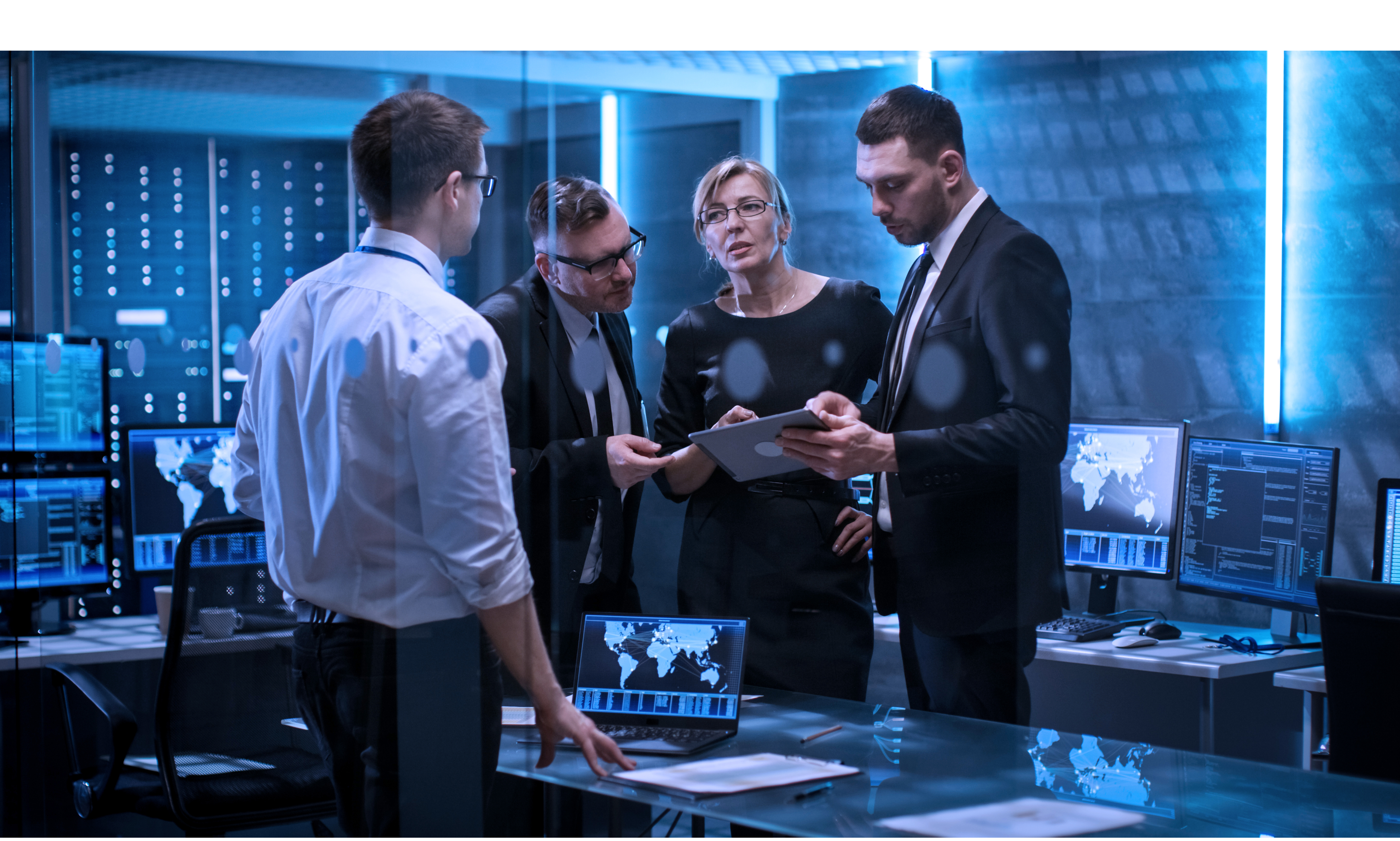 Photo of four technology professionals conferring as they look at a tablet in a modern office room that is illuminated with pale blue light