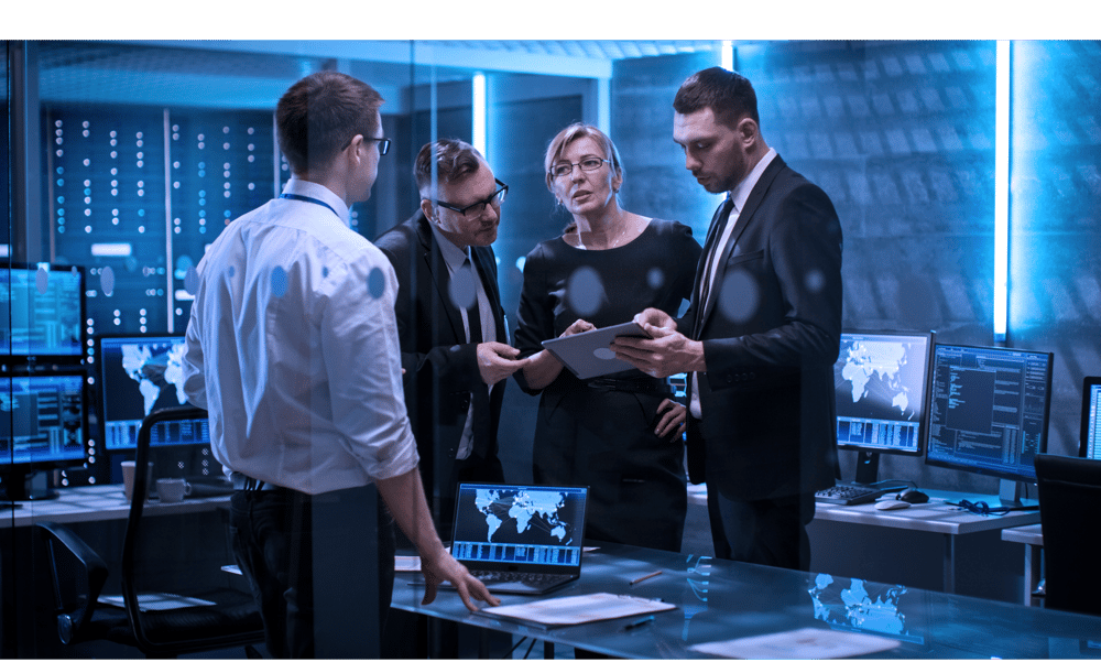 Photo of four technology professionals conferring as they look at a tablet in a modern office room that is illuminated with pale blue light