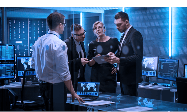 Photo of four technology professionals conferring as they look at a tablet in a modern office room that is illuminated with pale blue light Photo of four technology professionals conferring as they look at a tablet in a modern office room that is illuminated with pale blue light
