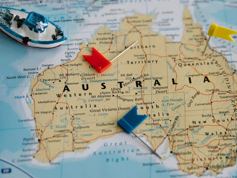 Mapa de Australia con banderas de colores y pequeños barcos de juguete sobre la superficie, señalando diferentes ubicaciones en el país, sobre un fondo de atlas.