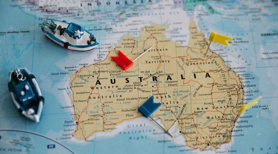 Mapa de Australia con banderas de colores y pequeños barcos de juguete sobre la superficie, señalando diferentes ubicaciones en el país, sobre un fondo de atlas.