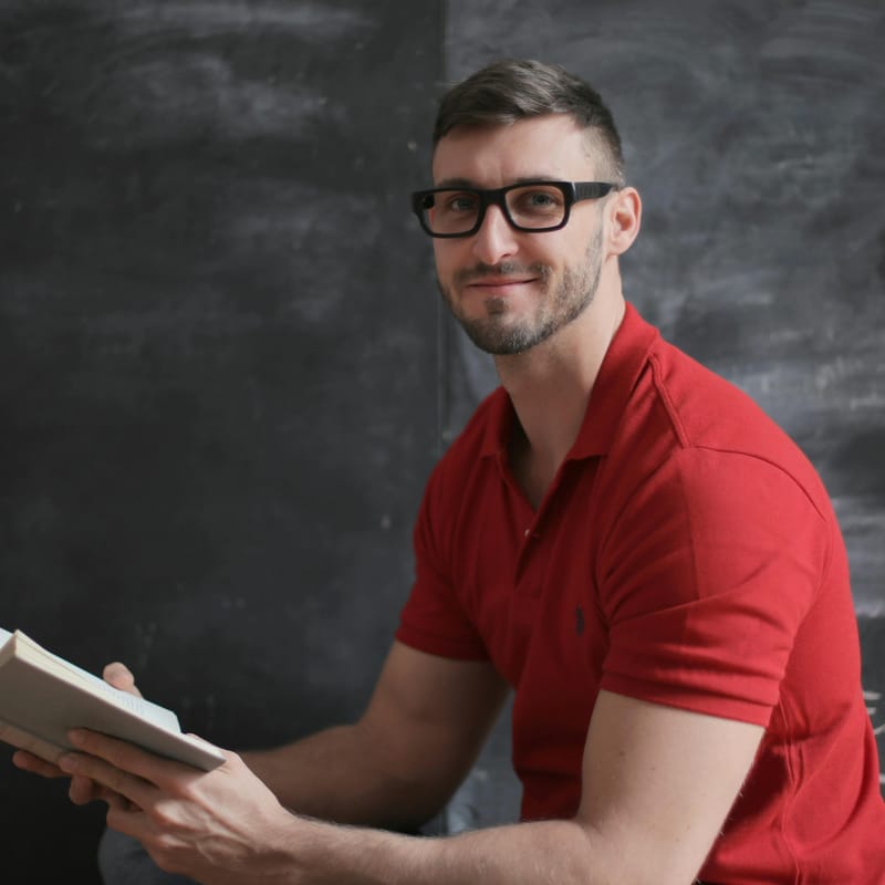 Hombre, leyendo un libro, sentado frente a un pizarrón.