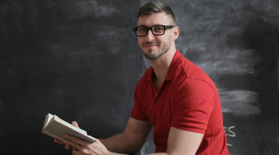 Hombre, leyendo un libro, sentado frente a un pizarrón.