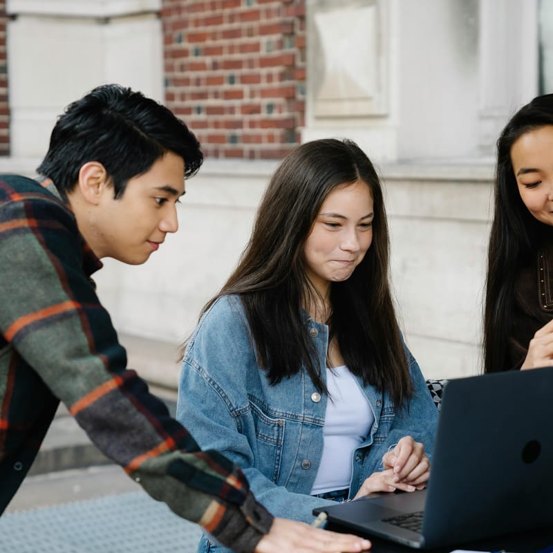 Tres estudiantes jóvenes colaboran frente a una laptop en un entorno universitario para estudiar temas de inglés c1.