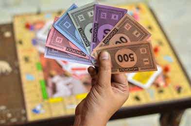 Hand holding Monopoly money in one hand with board game blurred in background.