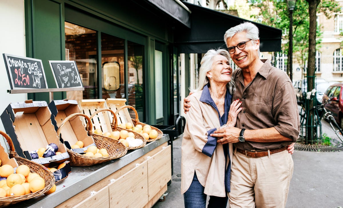 Una pareja de adultos mayores sonríe y se abraza frente a una tienda de frutas en la calle.