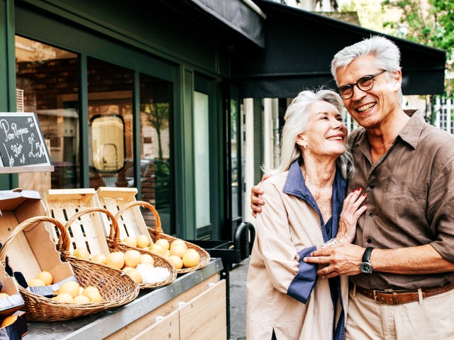 Una pareja de adultos mayores sonríe y se abraza frente a una tienda de frutas en la calle.