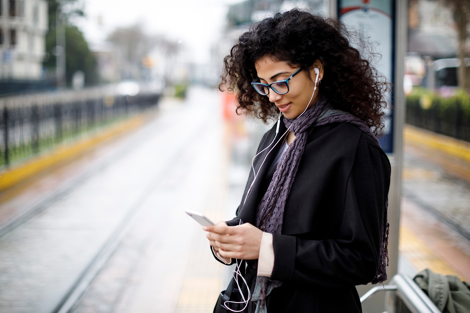 Una mujer con lentes y audífonos, usando su teléfono en la calle.