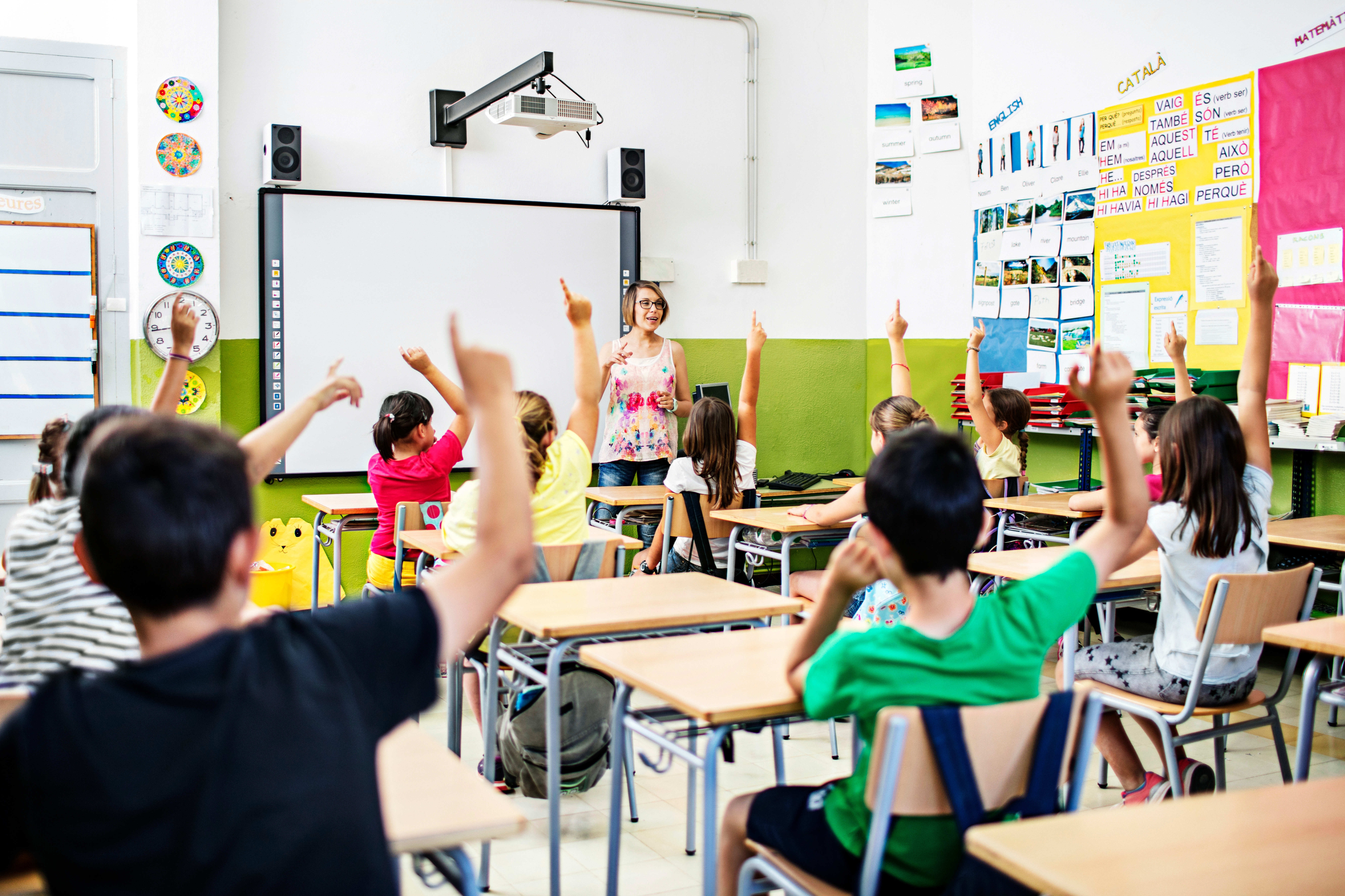 Una maestra interactúa con sus alumnos, quienes participan activamente levantando la mano en clase.