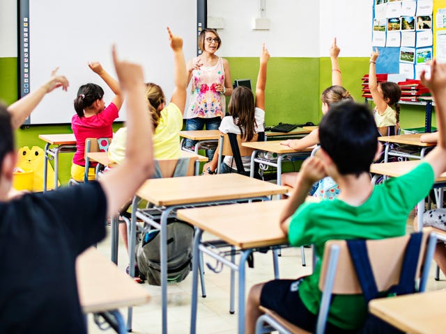 Una maestra interactúa con sus alumnos, quienes participan activamente levantando la mano en clase.