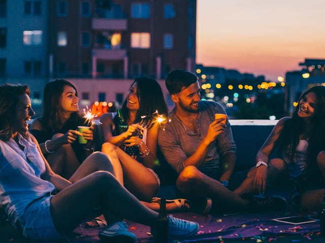 Un grupo de amigos ríe y celebra con bengalas en una azotea al atardecer o anochecer.