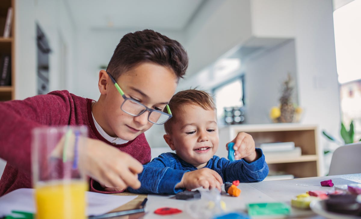 Dos hermanos, el mayor ayudando al más pequeño, juegan con plastilina en casa.