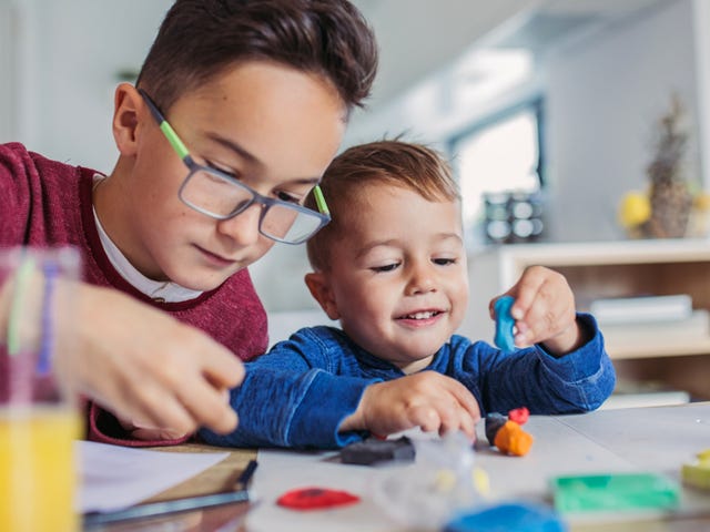 Dos hermanos, el mayor ayudando al más pequeño, juegan con plastilina en casa.