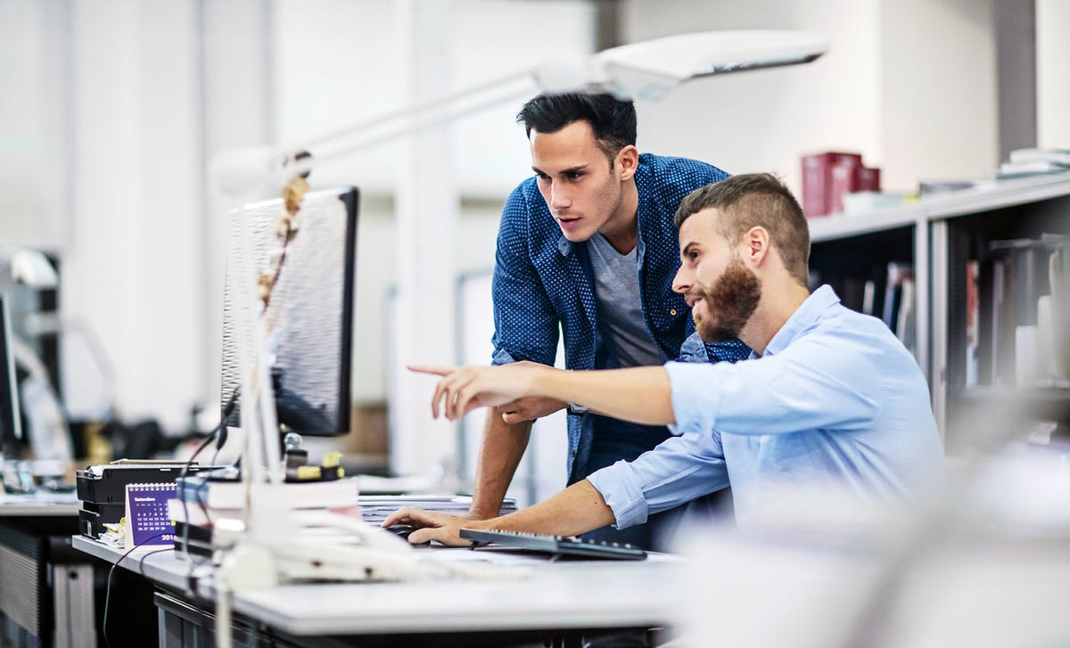 Dos hombres jóvenes colaboran y señalan una pantalla de computadora en un entorno de oficina.
