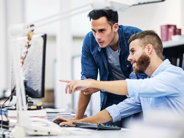 Dos hombres jóvenes colaboran y señalan una pantalla de computadora en un entorno de oficina.