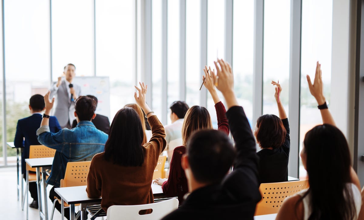 Un profesor se dirige a un grupo de personas que levantan la mano para participar.