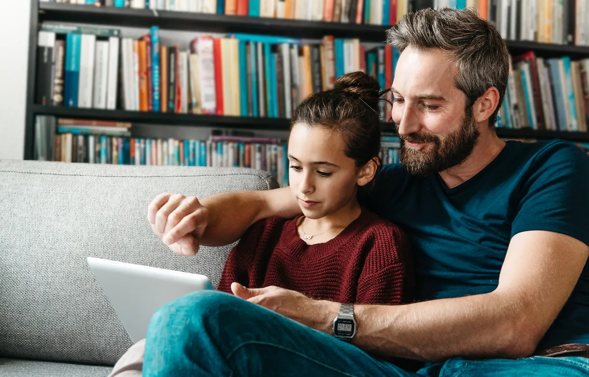 Un padre y su hija sentados juntos en el sofá, compartiendo un momento con una tablet.