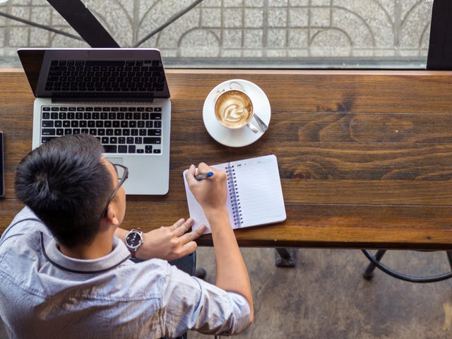 Un hombre joven concentrado, escribiendo en su libreta junto a su laptop y un café.