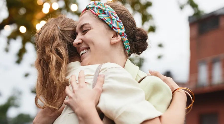 Mujer sonriendo mientras da un cálido abrazo a su amiga al aire libre.