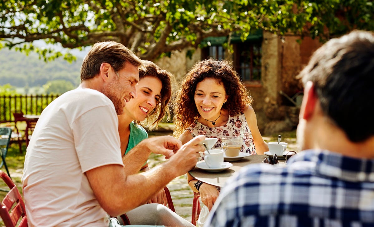 Un grupo de amigos sonríe mientras comparten un momento al aire libre, con tazas de café sobre la mesa.