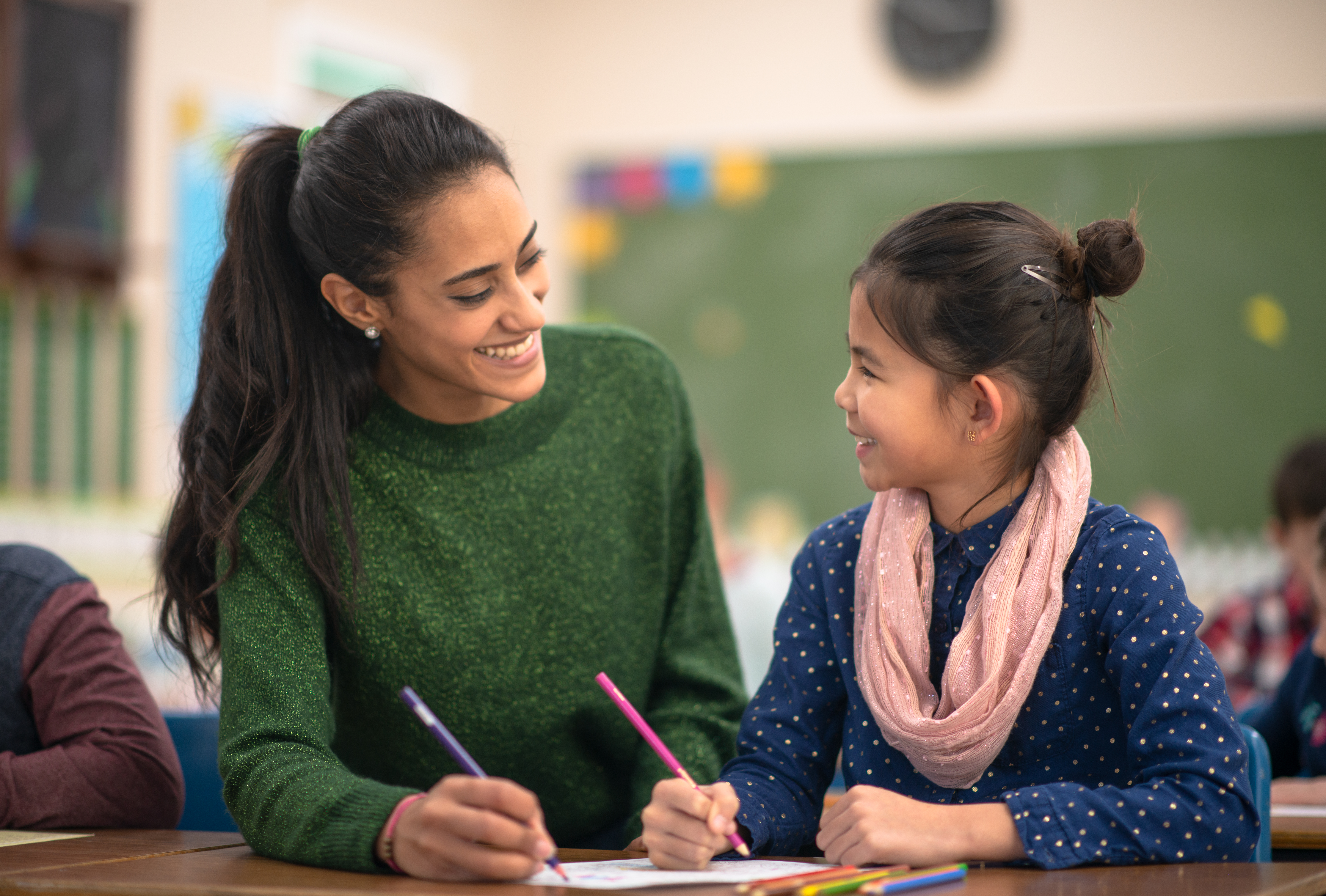 Una maestra joven guía y sonríe a una alumna, trabajando juntas en la clase.