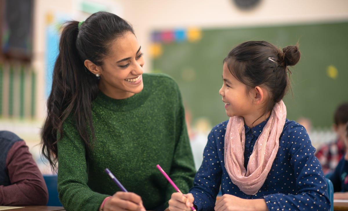 Una maestra joven guía y sonríe a una alumna, trabajando juntas en la clase.