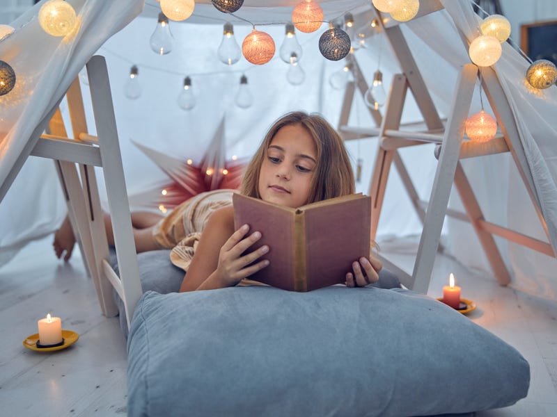 Niña leyendo libro en fuerte de mantas iluminado, aprendizaje creativo con Berlitz.